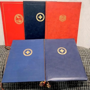 May include: A collection of vintage books in red and blue, featuring gold embossed logos and text. The books have a leather-like texture and are arranged on a soft, patterned surface. The logos include a cross within a circle.