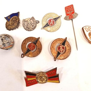 Könnte beinhalten: Eine Sammlung von Vintage-Anstecknadeln und -Abzeichen mit verschiedenen Designs und Farben. Einige Anstecknadeln haben Text, darunter "FDGB 25" und "DDR". Die Anstecknadeln zeigen eine Reihe von Formen, darunter Kreise, Schilde und eine Schleife. Die Farbpalette umfasst Gold, Rot, Schwarz und Blau.