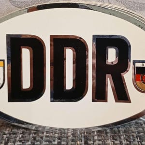Può includere: Adesivo ovale con le lettere "DDR" in nero. L'adesivo ha uno sfondo bianco e un bordo argentato. Piccoli emblemi con un orso e la bandiera della DDR su entrambi i lati delle lettere. Design in stile vintage.