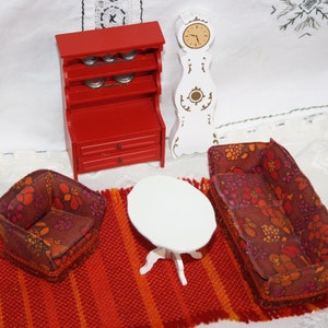 Könnte beinhalten: Ein Puppenhaus-Wohnzimmerset mit einer roten Anrichte, einer weißen Uhr, einem weißen Tisch und zwei floral gemusterten Sofas. Die Sofas und der Tisch stehen auf einem roten und orangefarbenen Teppich.