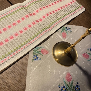 May include: A rectangular beige fabric runner featuring a repeating pattern of pink and green cross-stitched tulips and geometric lines, alongside a square beige fabric mat with cross-stitched pink tulips and blue flowers, topped by a brass candle holder.