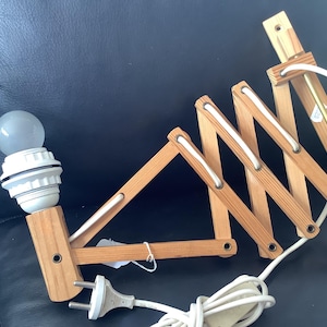 May include: A wooden wall lamp with a white cord and a white light bulb. The lamp has a folding design and can be adjusted to different angles.