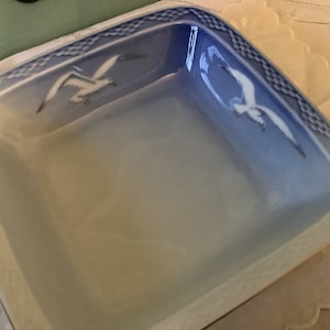 Puede incluir: Un plato cuadrado de cerámica blanca con un borde azul y un diseño de dos gaviotas en vuelo. El plato tiene un glaseado azul degradado sutil.