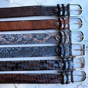 Peut inclure: Une collection de six ceintures présentant diverses textures et motifs, notamment des imprimés crocodile et peau de serpent dans des tons marron, gris et noir. Chaque ceinture est dotée d'une boucle en métal argenté et d'un passant assorti.