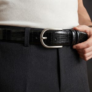 Könnte beinhalten: Ein schwarzer Gürtel mit glänzender Krokodilprägung und einer matten, silberfarbenen Metallschnalle. Der Gürtel ist durch eine Gürtelschlaufe gezogen und verfügt über einen Standard-Dornschließenverschluss mit einer strukturierten Optik.