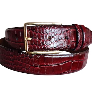May include: A burgundy leather belt with a gold-colored rectangular buckle. The belt has a textured, embossed pattern, and is rolled up. The buckle is a simple, classic design.