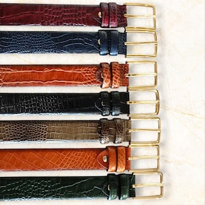 May include: A collection of seven leather belts in various colors, including burgundy, navy blue, brown, black, olive green, and orange. Each belt features a textured, embossed pattern and a gold-colored rectangular buckle.