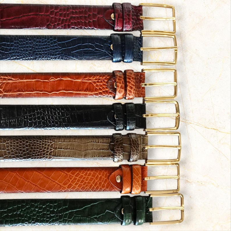 May include: A collection of seven leather belts in various colors, including burgundy, navy blue, brown, black, olive green, and orange. Each belt features a textured, embossed pattern and a gold-colored rectangular buckle.