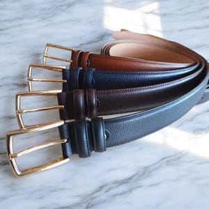 May include: A collection of four leather belts in tan, navy blue, dark brown, and black. Each belt features a textured surface and a polished gold-tone metal buckle with a rectangular frame and a single prong, arranged in a stacked, overlapping row.