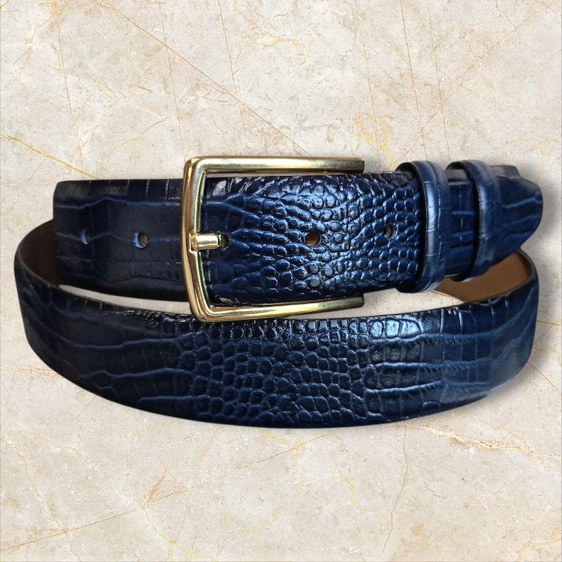 May include: A navy blue belt with a textured, crocodile-like pattern. The belt features a gold-colored rectangular buckle. The leather belt is a classic accessory.