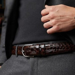 Op de afbeelding: Een donkerbruine riem met een glanzend, getextureerd krokodillenpatroon en een eenvoudige zilverkleurige metalen gesp. De riem wordt gedragen in een grijze broek, gecombineerd met een zwart bovenstuk en een donkergrijze blazer voor een formele stijl.