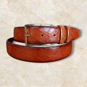 May include: A brown leather belt with a textured, crocodile-like pattern. The belt features a gold-colored rectangular buckle. The leather has varying shades of brown, creating a rich, detailed appearance. A classic accessory.