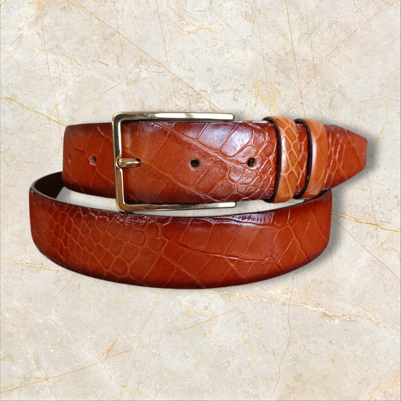 May include: A brown leather belt with a textured, crocodile-like pattern. The belt features a gold-colored rectangular buckle. The leather has varying shades of brown, creating a rich, detailed appearance. A classic accessory.