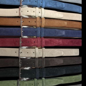 Op de afbeelding: Een verzameling suède riemen in diverse kleuren, waaronder grijs, blauw, beige, bruin, marineblauw, bordeauxrood, bruin, donkerbruin, zwart, groen en grijs. Elke riem heeft een zilverkleurige rechthoekige gesp en een bijpassende lus.