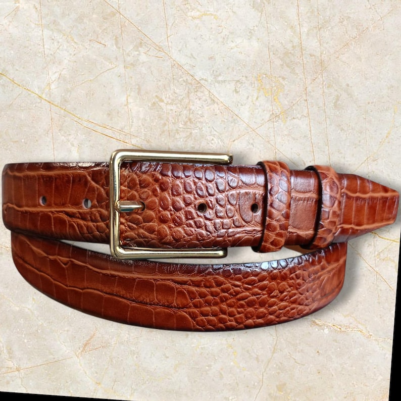 May include: A brown leather belt with a textured, crocodile-like pattern. The belt features a gold-colored rectangular buckle and a series of belt holes. The belt is a classic accessory.