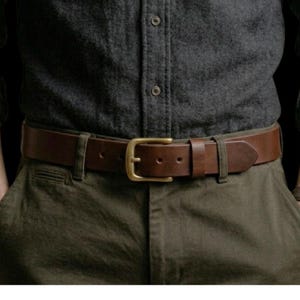 May include: A brown leather belt with a brass buckle is worn over olive green trousers. The belt is secured through the loops of the trousers. The person is wearing a dark grey button-down shirt.