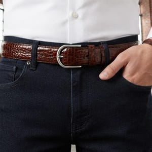 May include: A brown belt with a textured crocodile-skin pattern and a silver-toned metal buckle. The belt is worn through the loops of dark blue denim jeans, paired with a crisp white button-down shirt tucked into the waistband.