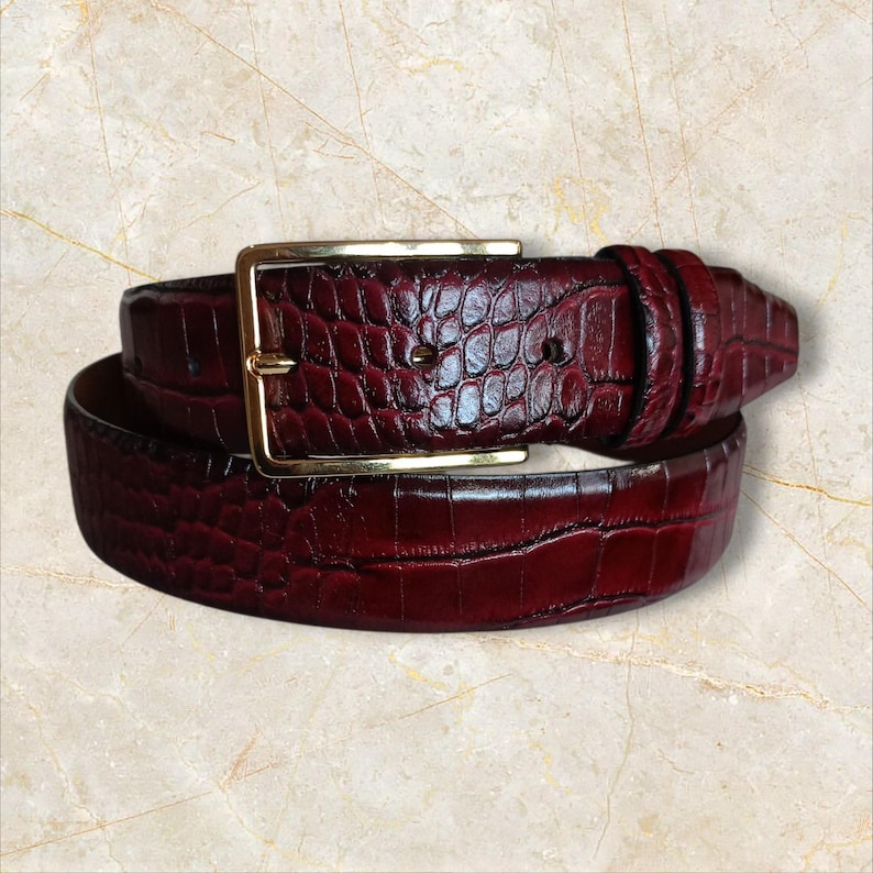 May include: A burgundy leather belt with a gold-colored rectangular buckle. The belt has a textured, crocodile-like pattern. The belt is rolled up, showing the buckle and the belt's texture.