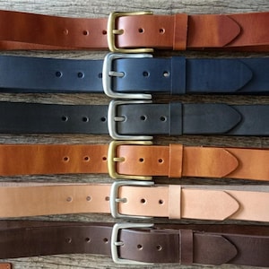 May include: Six leather belts in various colors, including brown, navy, and black, are displayed on a wooden surface. Each belt features a metal buckle, with some in silver and others in gold. The belts are of varying shades of brown.