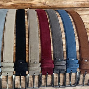 May include: A collection of suede belts in various colors, including light blue, tan, black, gray, burgundy, and brown. Each belt has a silver-colored buckle and a matching suede loop. The belts are arranged on a wooden surface.