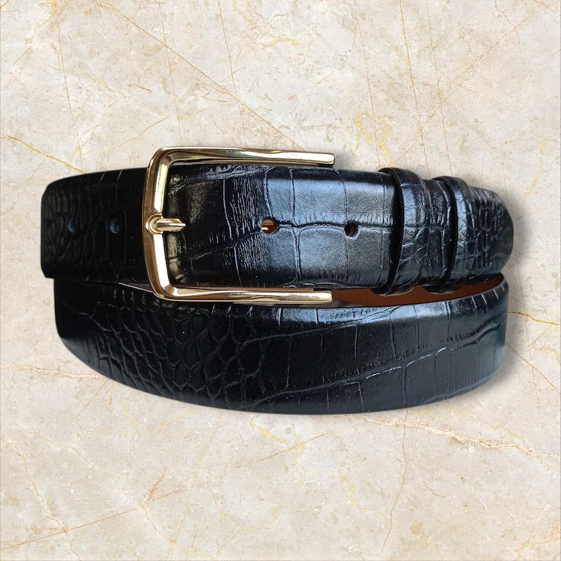 May include: A black belt with a gold-colored buckle. The belt has a textured, crocodile-like pattern. The buckle is rectangular and has a shiny finish. The belt is likely made of leather.