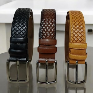 May include: Three leather belts in black, brown, and tan, each with a woven or textured pattern. The belts have silver-coloured metal buckles and loops. The black belt is on the left, the brown in the middle, and the tan belt is on the right.
