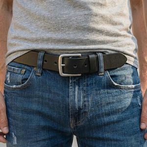 Pode incluir: Um cinto de couro preto com fivela prateada é usado com jeans azuis. O cinto é preso pelas presilhas dos jeans. A pessoa está vestindo uma camiseta cinza.