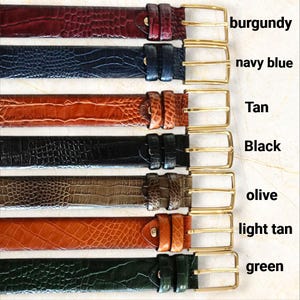 May include: A collection of seven leather belts in various colors, including burgundy, navy blue, tan, black, olive, light tan, and green. Each belt features a gold-colored buckle and a textured, crocodile-like pattern.