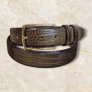 May include: A brown belt with a gold-colored rectangular buckle. The belt has a textured, crocodile-like pattern. The belt is rolled up on a light-colored surface.