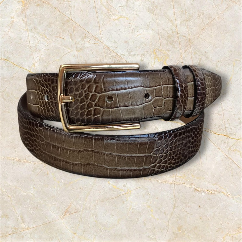 May include: A brown belt with a gold-colored rectangular buckle. The belt has a textured, crocodile-like pattern. The belt is rolled up on a light-colored surface.