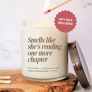 Puede incluir: Una vela blanca con una tapa marrón y una etiqueta que dice "Smells like she's reading one more chapter" y "The Flickery | Coconut Soy Wax | Vanilla Brown Sugar | 7.5 oz". La vela está sobre una superficie de madera con una cerilla encendida a su lado. Una pegatina roja en forma de estrella con el texto "Gift Box Included" está en la esquina superior derecha de la imagen.