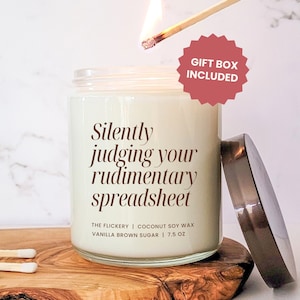 Peut inclure: Un bocal en verre contenant une bougie blanche avec un couvercle noir. La bougie est décorée de l'inscription "Silently judging your rudimentary spreadsheet". La bougie est étiquetée "The Flickery | Coconut Soy Wax | Vanilla Brown Sugar | 7.5 oz". Une allumette est en train d'être allumée pour allumer la bougie. Une étoile rouge avec le texte "Gift Box Included" est dans le coin supérieur droit de l'image.