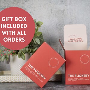 May include: Two red boxes with white text and a white circle logo. The boxes are for coconut soy wax candles. The text on the boxes reads "The Flickery" and "I was made just for you".