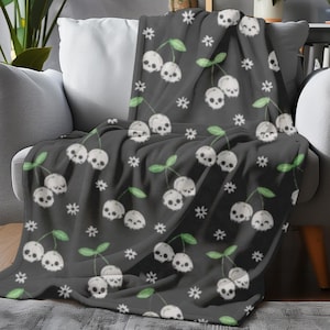 May include: A dark gray throw blanket featuring a repeating pattern of white skull cherries and small white star shapes. The blanket is draped over a gray armchair, creating a cozy and unique aesthetic.