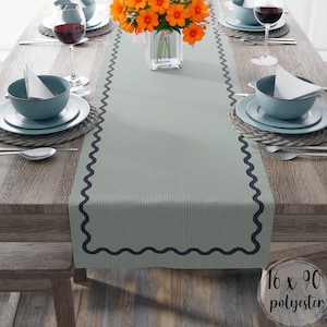 May include: A light green table runner with a small checkered pattern and a dark blue scalloped edge. The runner is 16 x 90 inches and made of polyester. The table is set with teal bowls, plates, and wine glasses.