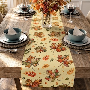May include: A cream-colored table runner featuring a repeating pattern of patchwork-style maple leaves in shades of orange, red, green, and brown. The runner is centered on a wooden table with place settings including bowls, plates, and glasses.