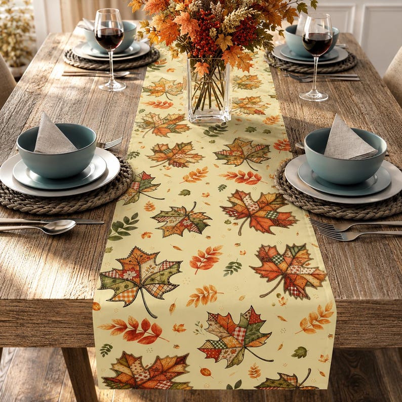 May include: A cream-coloured table runner featuring a repeating pattern of patchwork-style maple leaves in shades of orange, red, green, and brown. The runner is centred on a wooden table with place settings including bowls, plates, and glasses.