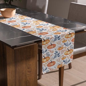 May include: A white table runner with a repeating pattern of orange pumpkins, blue leaves, and small orange berries. The runner is placed on a dark gray table, with a wooden table leg and a gray chair visible.