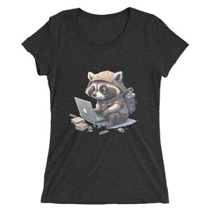 Puede incluir: Una camiseta de mujer en gris oscuro jaspeado con un dibujo de un mapache de dibujos animados que lleva una chaqueta con capucha y una mochila, usando un ordenador portátil. El mapache está rodeado de varios artículos de oficina, incluyendo un cuaderno, bolígrafos y una memoria USB.