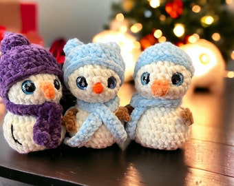 May include: Three crocheted plush snowmen figures made of white yarn. The left snowman wears a purple hat and scarf. The middle and right snowmen wear light blue hats and scarves. Each has large blue eyes, a small orange nose, and brown arms.