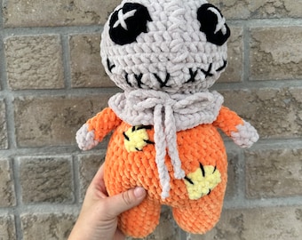 May include: A handmade crochet plush doll featuring a round beige head with black button eyes and a stitched mouth. The body is bright orange with yellow patches, and the doll wears a beige crocheted hood tied with a bow at the neck.