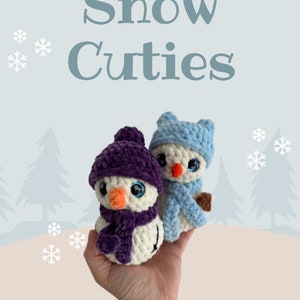 May include: Two small crocheted snowmen figures with plush white bodies and orange noses. One snowman wears a dark purple hat and scarf, while the other wears a light blue hat and scarf. The text Cassie Burchardt Snow Cuties is visible.