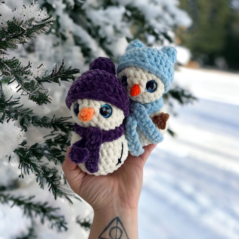 May include: Two small, handmade crochet snowman figures made from soft white yarn. One snowman wears a dark purple hat and scarf, while the other wears a light blue hat and scarf. Both have bright orange noses, large blue eyes, and round bodies.