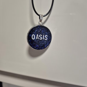 May include: A round pendant necklace with the word "OASIS" in white letters. The pendant has a dark blue, glittery background and a silver-colored metal border. The necklace has a black cord.