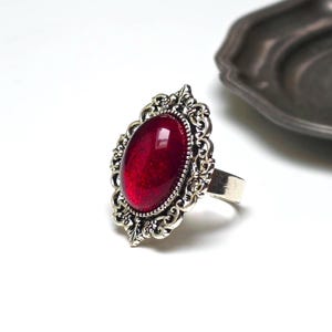 Op de afbeelding: Een zilverkleurige ring met een ovale, dieprode cabochonsteen in een gedetailleerde, filigraanachtige metalen zetting. De ringband is effen en van metaal, wat een contrast vormt met de sierlijke, decoratieve omlijsting rondom de centrale steen.