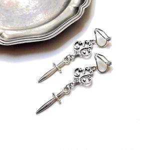 May include: A pair of silver-toned earrings with a key and dagger design. The earrings feature a decorative scrollwork pattern and a white, petal-shaped accent. The earrings are displayed on a white surface near a silver-colored decorative plate.