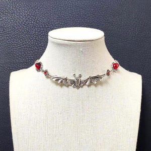 Op de afbeelding: Een zilverkleurige metalen choker-halsketting met een centrale vleermuishanger met uitgestrekte vleugels. De ketting is versierd met kleine, ronde, rode gefacetteerde stenen in zilveren zettingen, wat zorgt voor een symmetrisch ontwerp rond de hals.