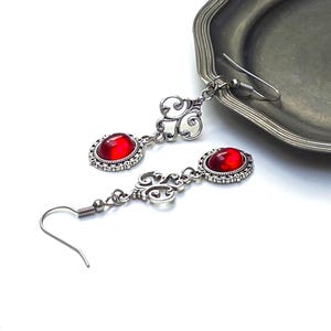 Op de afbeelding: Zilverkleurige oorhangers met ovale, dieprode edelstenen. De oorbellen hebben een sierlijk filigraan ontwerp en een haaksluiting. De oorbellen worden gepresenteerd op een wit oppervlak naast een zilveren dienblad.