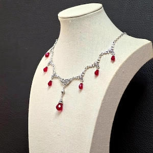 Op de afbeelding: Een zilverkleurige metalen ketting met gedetailleerd filigraanwerk en meerdere bungelende rode druppelvormige kralen. De ketting bestaat uit fijne schakels, waarbij de rode kralen met tussenpozen langs de voorzijde van het sieraad hangen.