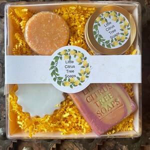 May include: A gift box filled with yellow shredded paper and three lemon-themed bath products. The box contains a round bar of soap, a round container of soap, and a rectangular bar of soap. The labels on the products read "Little Citrus Tree Soaps".